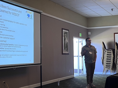 A man gestures while speaking beside a projector screen.